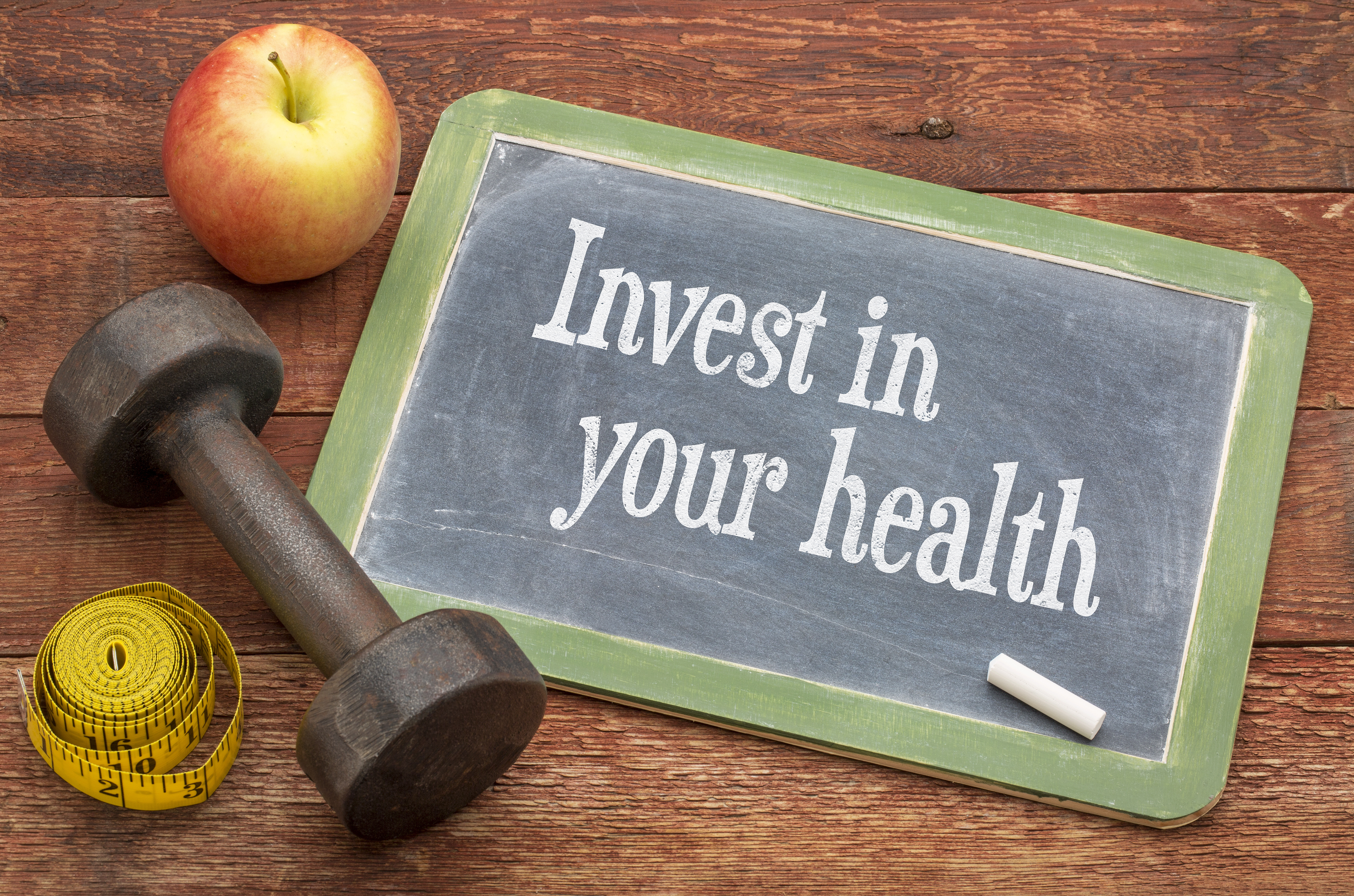 Invest in your health - slate blackboard sign against weathered red painted barn wood with a dumbbell, apple and tape measure